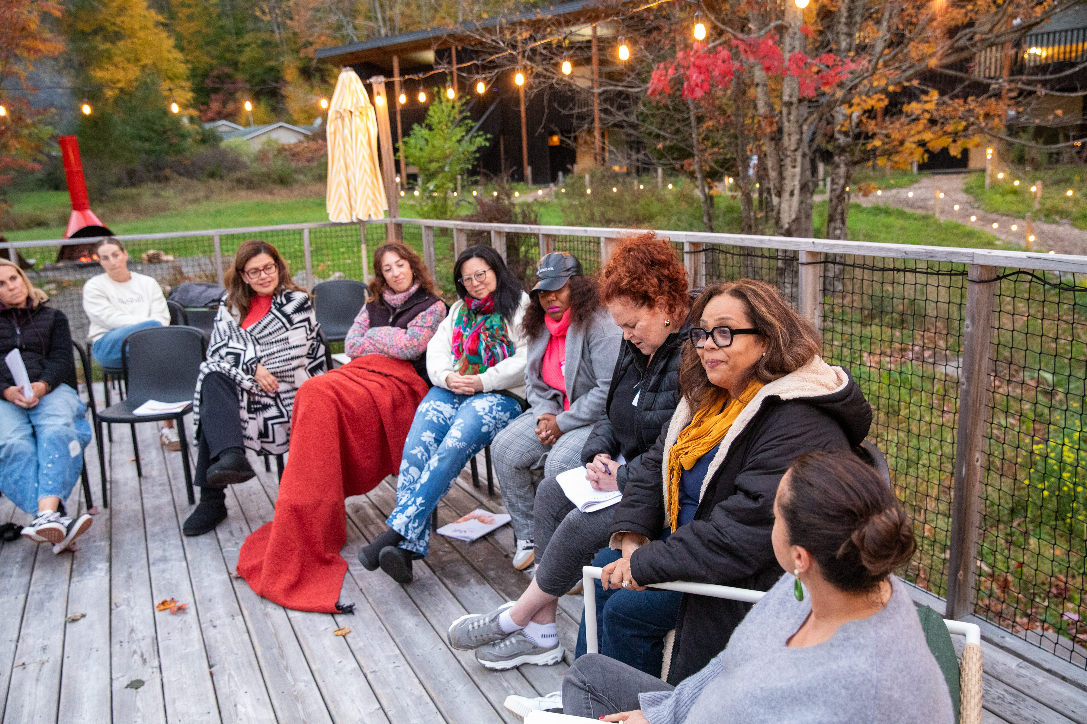 Women gathered in conversation on the deck beneath string lights and autumn trees at The Shift